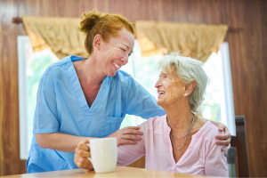 Home care nurse smiling with hands on shoulders of older woman sitting at table smiling and drinking tea